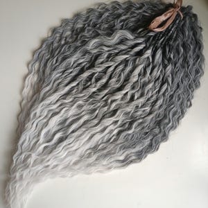 Könnte beinhalten: Ein Set aus synthetischen Haarverlängerungen mit welliger, texturierter Struktur und einem Ombré-Farbverlauf, der von dunkelanthrazitfarbenen Ansätzen zu hellem Silber und Weiß an den Spitzen übergeht, zusammengehalten durch ein hellbraunes Band.