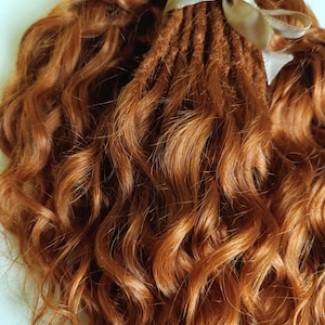 Puede incluir: Un conjunto de rastas de color castaño rojizo con cabello suelto y ondulado. Las rastas están atadas con una cinta marrón claro. El cabello tiene una apariencia natural y texturizada.