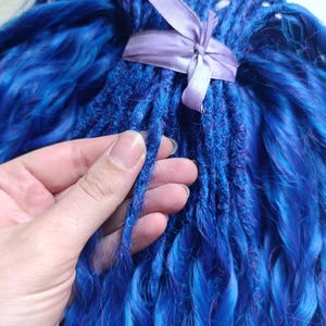 May include: A collection of bright blue synthetic dreadlocks, secured with a lavender ribbon. The dreadlocks display a blend of deep and lighter blue tones, with a hand holding some strands, highlighting the texture and colour variations. The overall appearance is striking and vibrant.