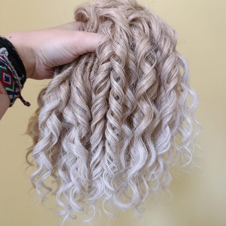 May include: A bundle of blonde and white curly hair extensions. The hair is held in a hand, showcasing the texture and color gradient from blonde to white. The curls are tightly wound and voluminous.