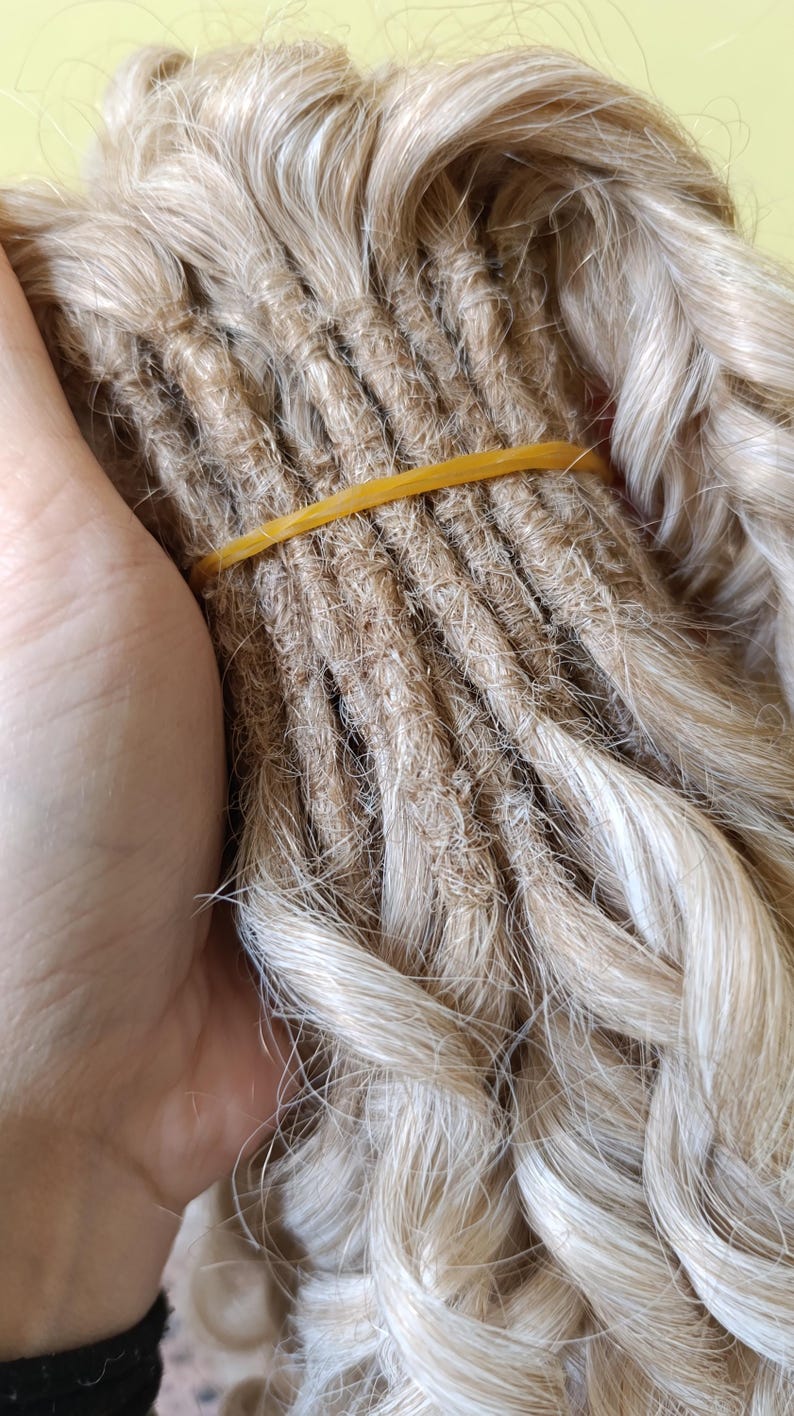 May include: Close-up of a bundle of blonde dreadlocks, secured with a yellow rubber band. The dreadlocks have a textured, natural appearance, with varying shades of blonde and brown. The hair is styled in a twisted, rope-like fashion.