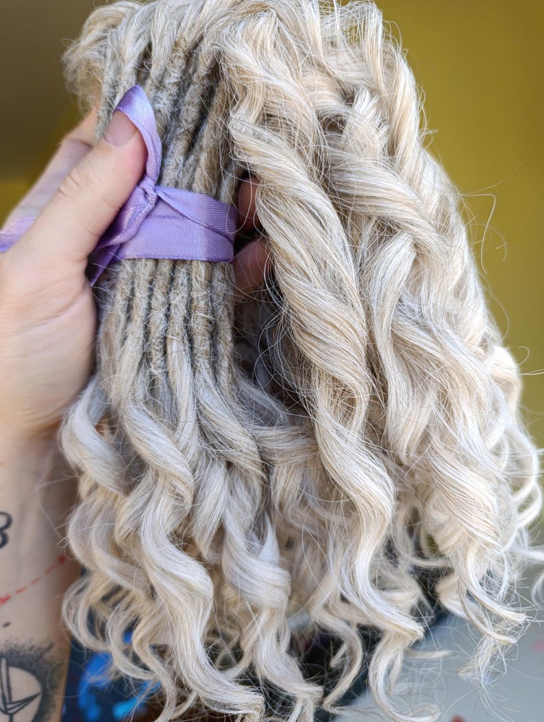 May include: A bundle of light gray dreadlocks with curly ends, secured with a lavender ribbon. The dreadlocks have a natural, textured appearance, and the curls add volume and style. The overall look is soft and textured.