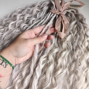 Op de afbeelding: Een close-up van een lichtblond krullend haarstuk met een bruin lint dat bovenaan is vastgebonden. Het haar is lang en dik, en het lint is in een strik geknoopt.