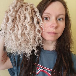 May include: A bundle of light blonde, curly hair extensions. The hair is held up against a person's face, with the person wearing a blue t-shirt. The background is a solid yellow color.