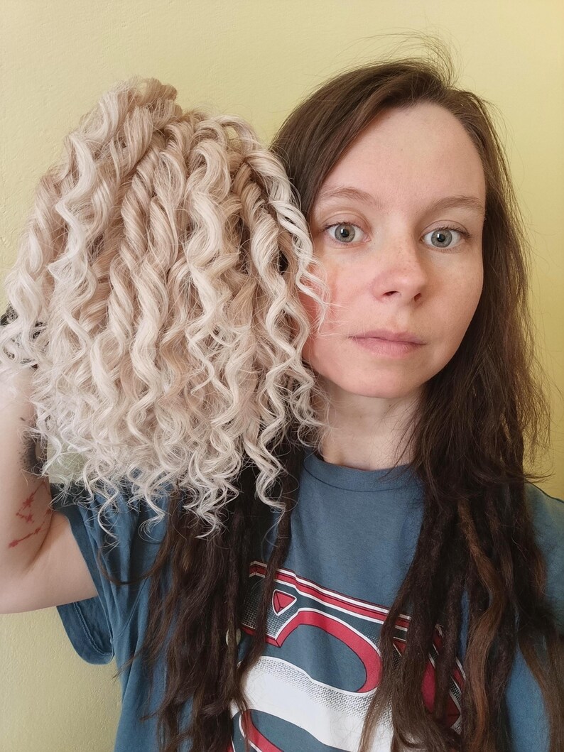 May include: A bundle of light blonde, curly hair extensions. The hair is held up against a person's face, with the person wearing a blue t-shirt. The background is a solid yellow color.