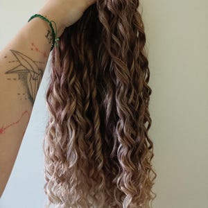 May include: A bundle of long, curly hair extensions in shades of brown and blonde. The hair is held up against a plain background, showcasing the texture and color variations. The curls are tightly wound and cascade downwards.