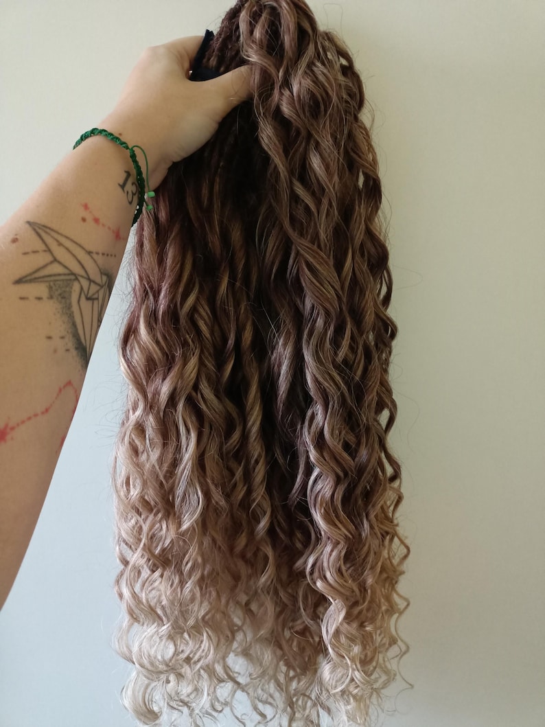 May include: A bundle of long, curly hair extensions in shades of brown and blonde. The hair is held up against a plain background, showcasing the texture and color variations. The curls are tightly wound and cascade downwards.