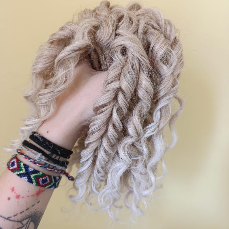 May include: A light blonde wig with tight, spiral curls. The wig is held in a hand, showcasing the texture and style of the hair. The curls are a mix of light blonde and grey tones.