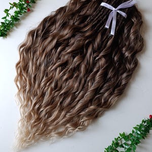 May include: A bundle of brown and blonde wavy hair extensions tied with a white ribbon. The hair has a natural gradient, transitioning from dark brown at the roots to a lighter blonde at the ends. The extensions are styled with loose, voluminous waves.