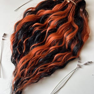 May include: A bundle of wavy, synthetic hair extensions in black and vibrant orange. The hair is styled with a mix of dreadlocks and loose waves, secured with a light brown ribbon. Dried lavender sprigs are arranged around the hair on a white surface.