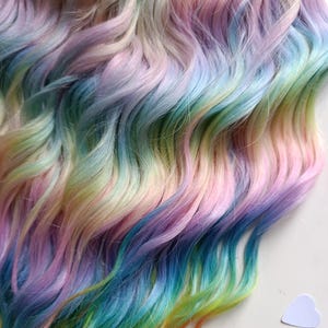 May include: A close-up of wavy, multi-coloured hair extensions in pastel shades of pink, blue, purple, yellow, and green. The hair is arranged in flowing, layered strands, creating a vibrant rainbow effect. A small, white heart-shaped object is in the lower right corner.