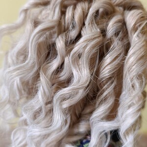 May include: Close-up of a head of light blonde, curly hair. The curls are tightly wound and have a natural, textured appearance. The hair is styled with a slight wave, and the color is a mix of blonde and light brown.