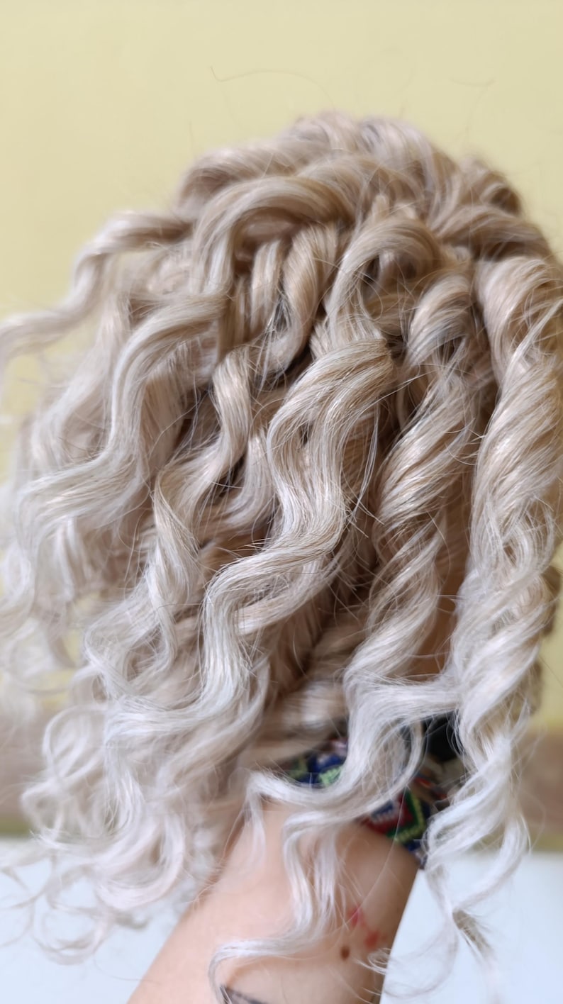 May include: Close-up of a head of light blonde, curly hair. The curls are tightly wound and have a natural, textured appearance. The hair is styled with a slight wave, and the color is a mix of blonde and light brown.
