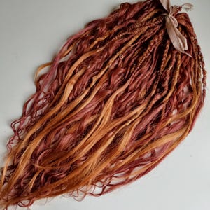 Peut inclure: Ensemble de dreadlocks roux et orange clair avec une texture ondulée. Les dreadlocks sont attachés avec un ruban marron clair. Les extensions de cheveux sont disposées sur une surface blanche.