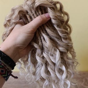 May include: A bundle of blonde and light brown curly hair extensions. The extensions are held in a hand, showcasing the texture and color variations. The curls are tightly wound and create a voluminous look.