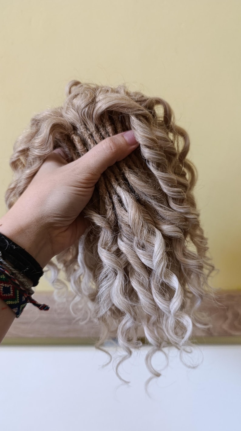 May include: A bundle of blonde and light brown curly hair extensions. The extensions are held in a hand, showcasing the texture and color variations. The curls are tightly wound and create a voluminous look.