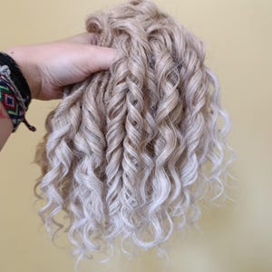 May include: A bundle of light blonde curly hair extensions with a gradient effect. The hair is held in a hand, showcasing the texture and color variations. The curls are tightly wound and cascade downwards.