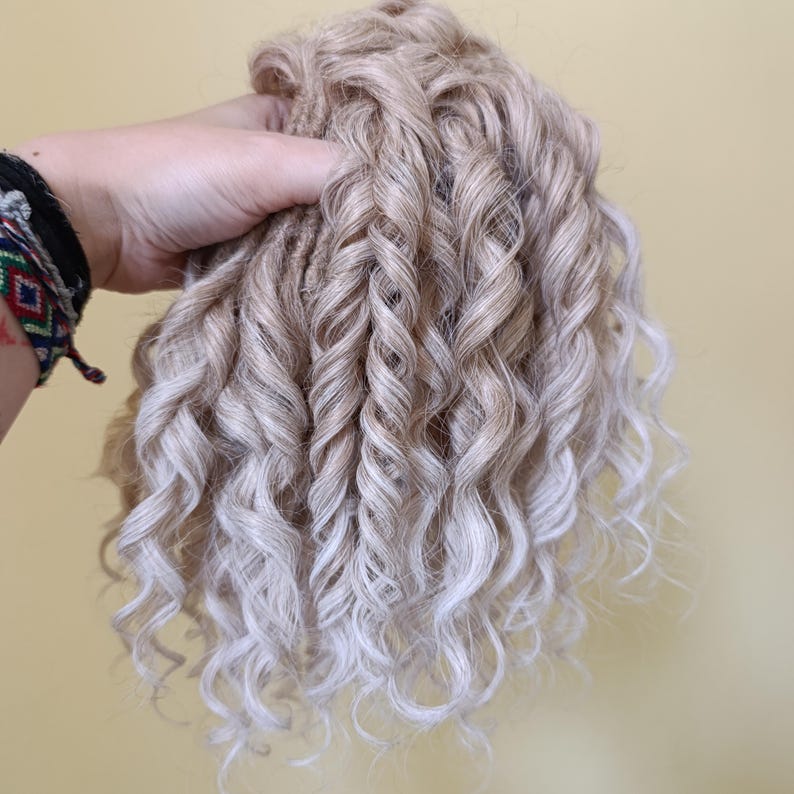 May include: A bundle of light blonde curly hair extensions with a gradient effect. The hair is held in a hand, showcasing the texture and color variations. The curls are tightly wound and cascade downwards.