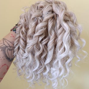 May include: A close-up of a bundle of blonde curly hair with highlights. The hair is styled in tight, voluminous curls, with a mix of light and darker blonde tones. The curls cascade in a natural, flowing pattern.