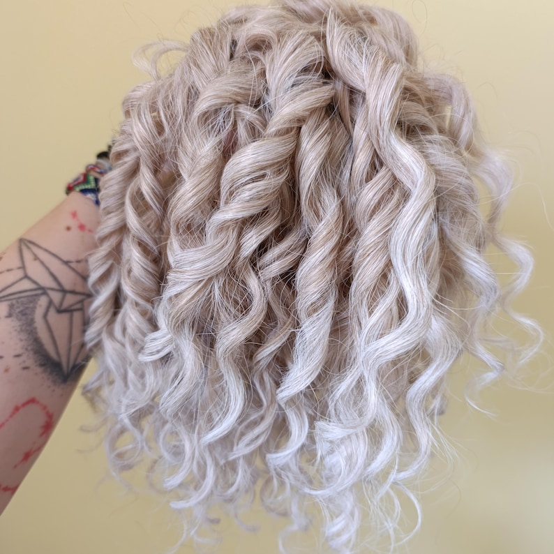 May include: A close-up of a bundle of blonde curly hair with highlights. The hair is styled in tight, voluminous curls, with a mix of light and darker blonde tones. The curls cascade in a natural, flowing pattern.