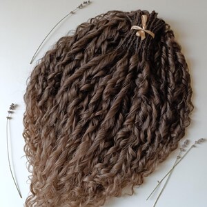 May include: A bundle of brown and light brown curly hair extensions. The hair is styled in tight, voluminous curls, with a darker root and lighter tips. A gold ribbon secures the top of the bundle. Sprigs of lavender are placed around the hair.