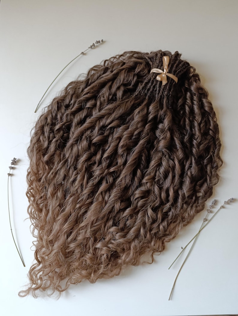 May include: A bundle of brown and light brown curly hair extensions. The hair is styled in tight, voluminous curls, with a darker root and lighter tips. A gold ribbon secures the top of the bundle. Sprigs of lavender are placed around the hair.