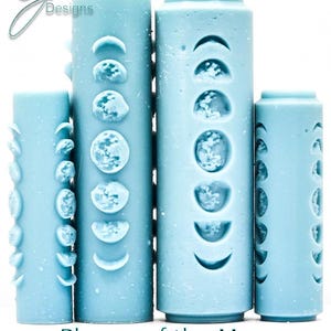 May include: Four light blue pillar candles featuring moon phase designs. The candles are cylindrical and of varying heights. The text "Phases of the Moon" and "Deepbression" is at the bottom. The candles are set against a white backdrop.
