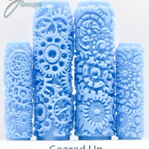 May include: Four light blue cylindrical rollers with intricate gear and floral patterns. The rollers are arranged in a row, showcasing a variety of detailed designs. The text "Geared Up Deep Texture" is displayed below the rollers.