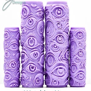 May include: Four cylindrical purple paint rollers featuring a raised, embossed pattern of numerous realistic eyes. The rollers vary in height, with two taller central rollers and two shorter outer rollers. The text "Watching You Intricate Texture" is printed below the rollers, and a "G Designs" logo is visible in the top left corner.