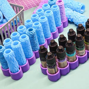 May include: An assortment of blue and purple patterned texture rollers and small bottles of stain. The rollers are in a gray basket and arranged in rows. The bottles are in purple holders.