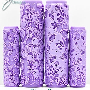 Puede incluir: Cuatro rodillos de amasar de color lavanda con intrincados diseños de abejas, panales y flores. El texto "Bizzy Bees Intricate Texture" se muestra debajo. Los rodillos varían en tamaño y están dispuestos verticalmente.