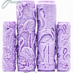 Könnte beinhalten: Mehrere lavendelfarbene Säulenkerzen mit aufwendigen Flamingodesigns. Die Kerzen zeigen detaillierte Schnitzereien von Flamingos, tropischen Pflanzen und den Text "Flamingos Intricate Texture". Die Kerzen sind unterschiedlich hoch.