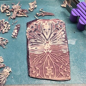 May include: A close-up of a patterned clay tile with a detailed design in shades of brown and cream. The tile is on a blue surface with pieces of broken clay around it.