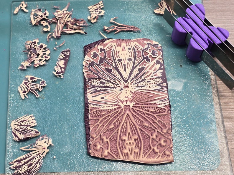 May include: A close-up of a patterned clay tile with a detailed design in shades of brown and cream. The tile is on a blue surface with pieces of broken clay around it.