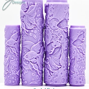 May include: Four cylindrical lavender paint rollers featuring embossed goldfish and bubble patterns. The rollers display the text G Designs, Goldfish, and Intricate Texture. Each roller has a raised surface design for creating textured patterns.