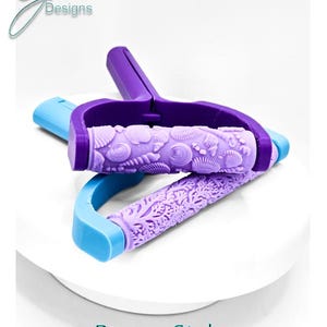 May include: Two brayer style rollers with textured purple cylinders. One roller features a seashell pattern, while the other displays a coral reef design. The handles are purple and light blue. The text G Designs and Brayer Style RollerRoller are visible.