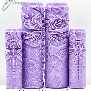 Könnte beinhalten: Vier lavendelfarbene Zylinder mit geätzten Libellen- und Blattmustern. Die Objekte sind unterschiedlich groß und in einer Reihe angeordnet. Der Text "Etched Dragonfly Intricate Texture" befindet sich unten. Detaillierte, strukturierte Oberfläche.