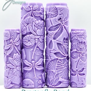 May include: Four lavender-coloured, cylindrical textured rollers with dragonfly and lotus flower designs. The rollers are of varying heights and feature the text "Dragonfly Pond Intricate Texture". The rollers are for crafting or decorative purposes.