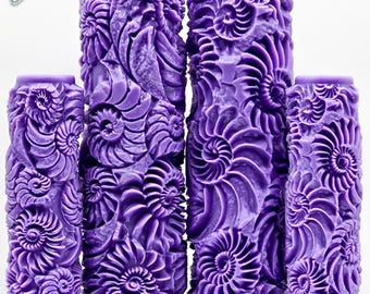 May include: Four purple patterned paint rollers featuring a raised, embossed design of fossilized nautilus shells. The rollers vary in height and width, displaying an intricate, repeating spiral texture across their entire cylindrical surfaces.