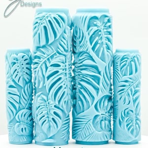 May include: Four light blue cylindrical paint rollers featuring a raised Monstera leaf pattern. The rollers vary in height, with two taller ones in the center and two shorter ones on the sides. The text G Designs, Monstera, and Deep Impression is visible.