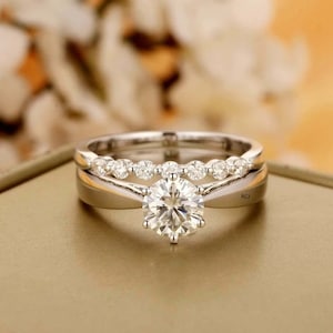 Puede incluir: Un juego de alianzas de boda de plata con una piedra central grande, redonda y transparente y una banda más pequeña de piedras más pequeñas y transparentes.