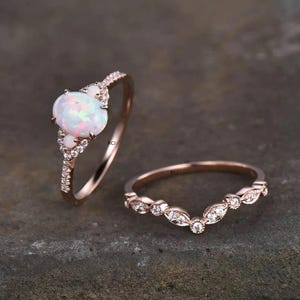 May include: Two rose gold rings. One ring features a large oval opal gemstone with smaller accent stones. The second ring is a curved band with small, clear gemstones. Both rings are on a dark gray surface.