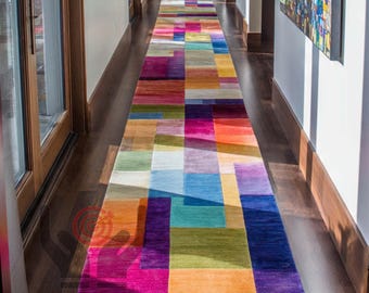 May include: A long, rectangular area rug with a geometric pattern of colorful blocks. The rug features a vibrant mix of colors including purple, orange, yellow, green, blue, and pink. The rug is placed in a hallway with dark wood flooring.