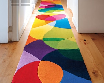 Pode incluir: Um tapete comprido e colorido com um design abstrato de círculos sobrepostos em tons de vermelho, laranja, amarelo, verde, azul, roxo e rosa. O tapete está colocado sobre um chão de madeira num corredor.