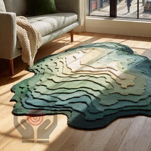 May include: A distinctive rug with a topographical design, featuring a layered effect in shades of green, blue, and beige. The rug's design creates a textured, three-dimensional appearance. It is placed on a wooden floor near a grey sofa.