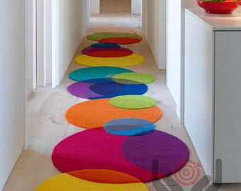 Pode incluir: Um tapete de corredor composto por tapetes circulares coloridos sobrepostos. Os tapetes são em tons de amarelo, verde, azul, roxo, laranja e rosa. O tapete é colocado sobre um chão de madeira clara.