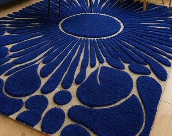 Pode incluir: Um tapete grande e quadrado com um impressionante design floral azul. O tapete apresenta um círculo azul central rodeado por formas azuis em forma de pétalas radiantes sobre um fundo bege neutro. O tapete está colocado sobre um chão de madeira.