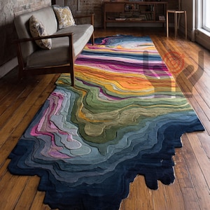 Könnte beinhalten: Ein farbenfroher Teppich mit topografischem Design. Der Teppich weist einen Farbverlauf aus Blau-, Grün-, Gelb-, Orange- und Rosatönen auf, wodurch ein geschichteter Effekt entsteht. Der Teppich liegt auf einem Holzboden, im Hintergrund ein Sofa und eine Backsteinmauer.