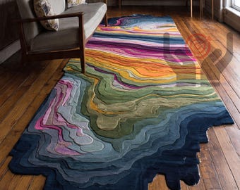 May include: A vibrant, multi-coloured rug with a topographical design. The rug's design features a gradient of blues, greens, yellows, oranges, and pinks, creating a layered effect. The rug is placed on a wooden floor, with a sofa and brick wall in the background.