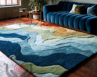 May include: A large, rectangular area rug with a layered, abstract design in shades of blue, green, and tan. The rug is placed on a hardwood floor, with a blue sofa and small side tables in the background. The rug's design resembles a topographical map.